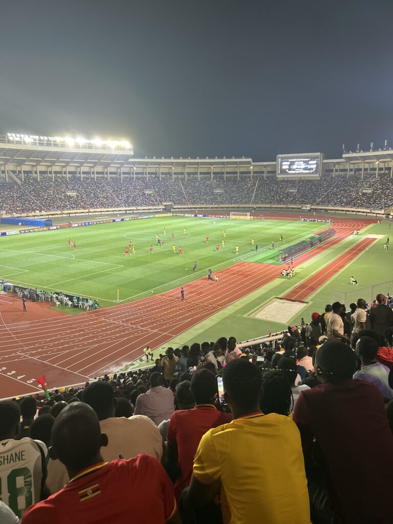 Football supporters overlook the floodlit pitch and running track at Namboole Stadium.